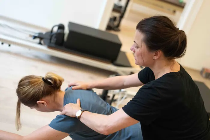 Kézzel végzett lapocka-korrekció ülő helyzetben, a háttérben Pilates reformer gép.